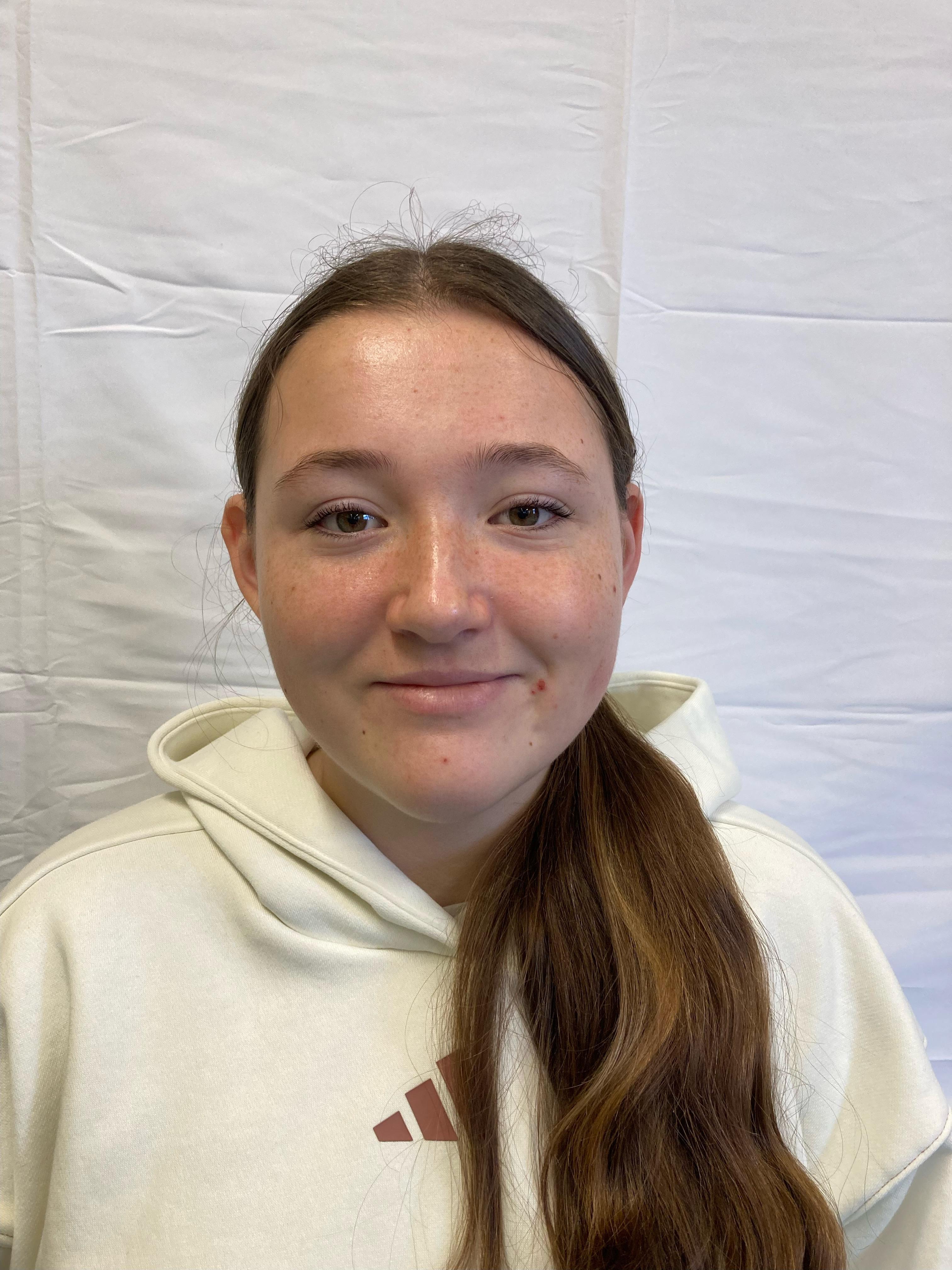 A girl with tied-up brunette hair wearing a white hoodie poses for a profile picture.