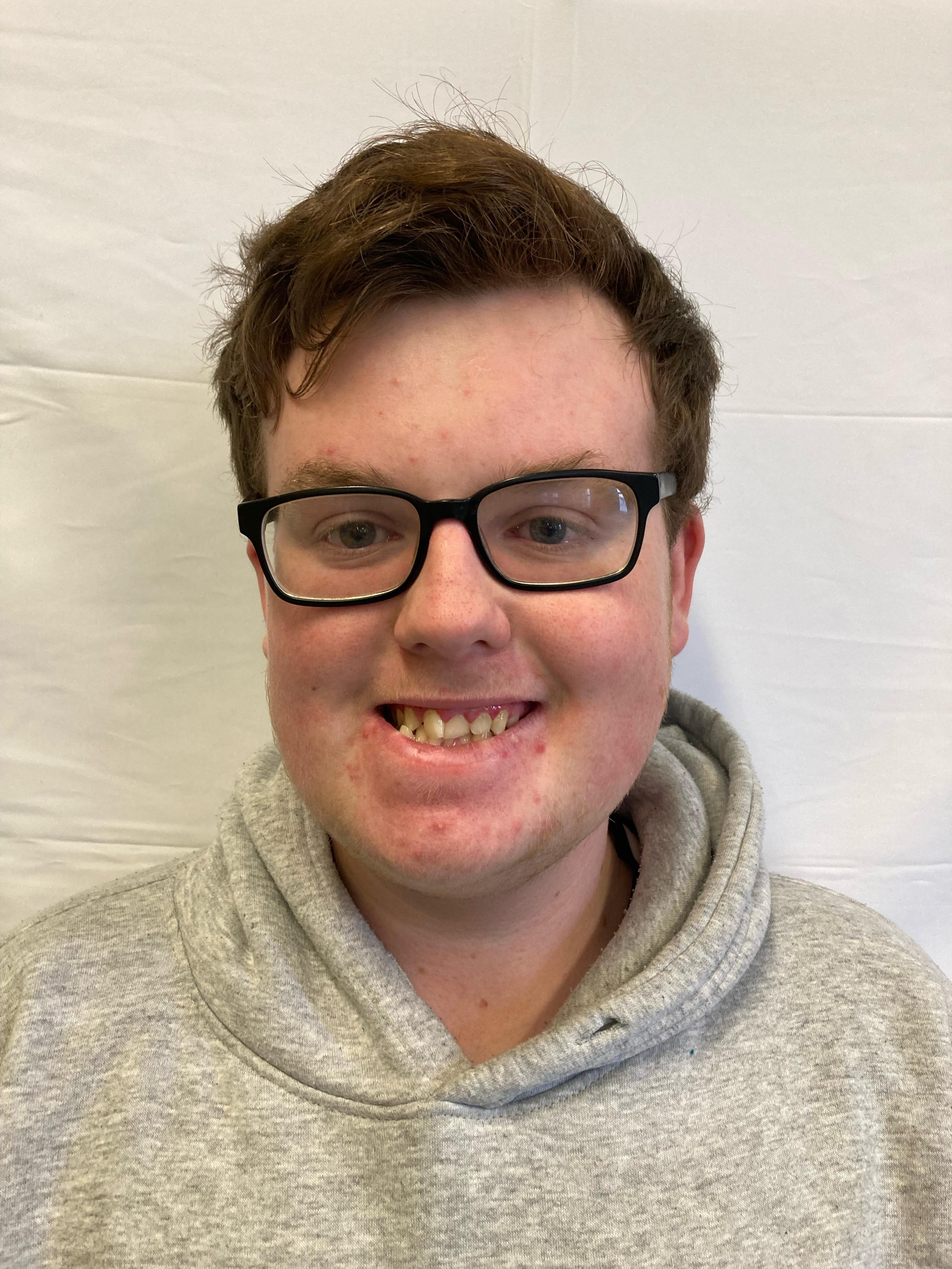 A boy with brunette hair wearing glasses and a grey hoodie poses for a profile picture.