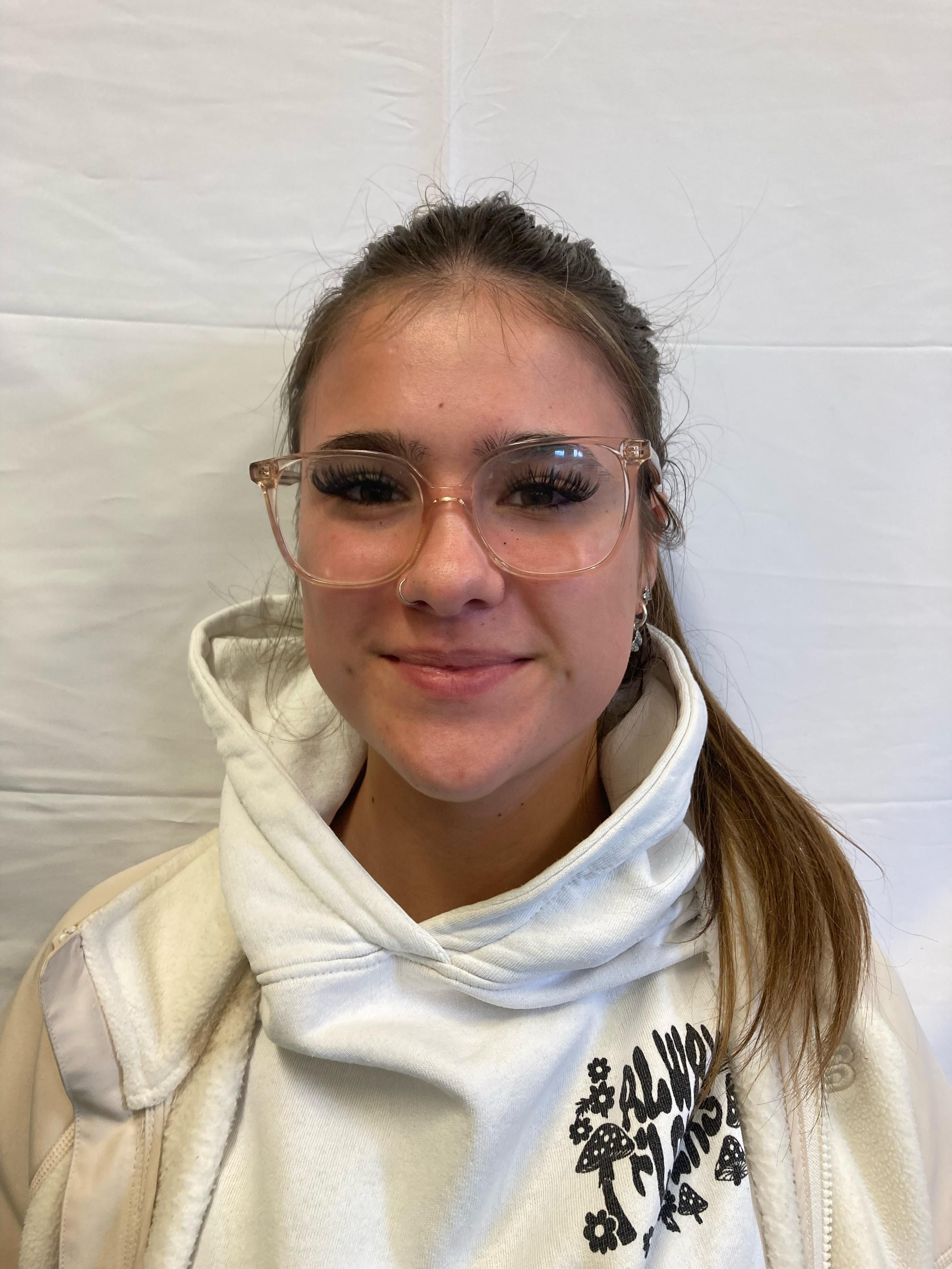 A girl with tied-up brunette hair wearing glasses and a white hoodie poses for a profile picture.