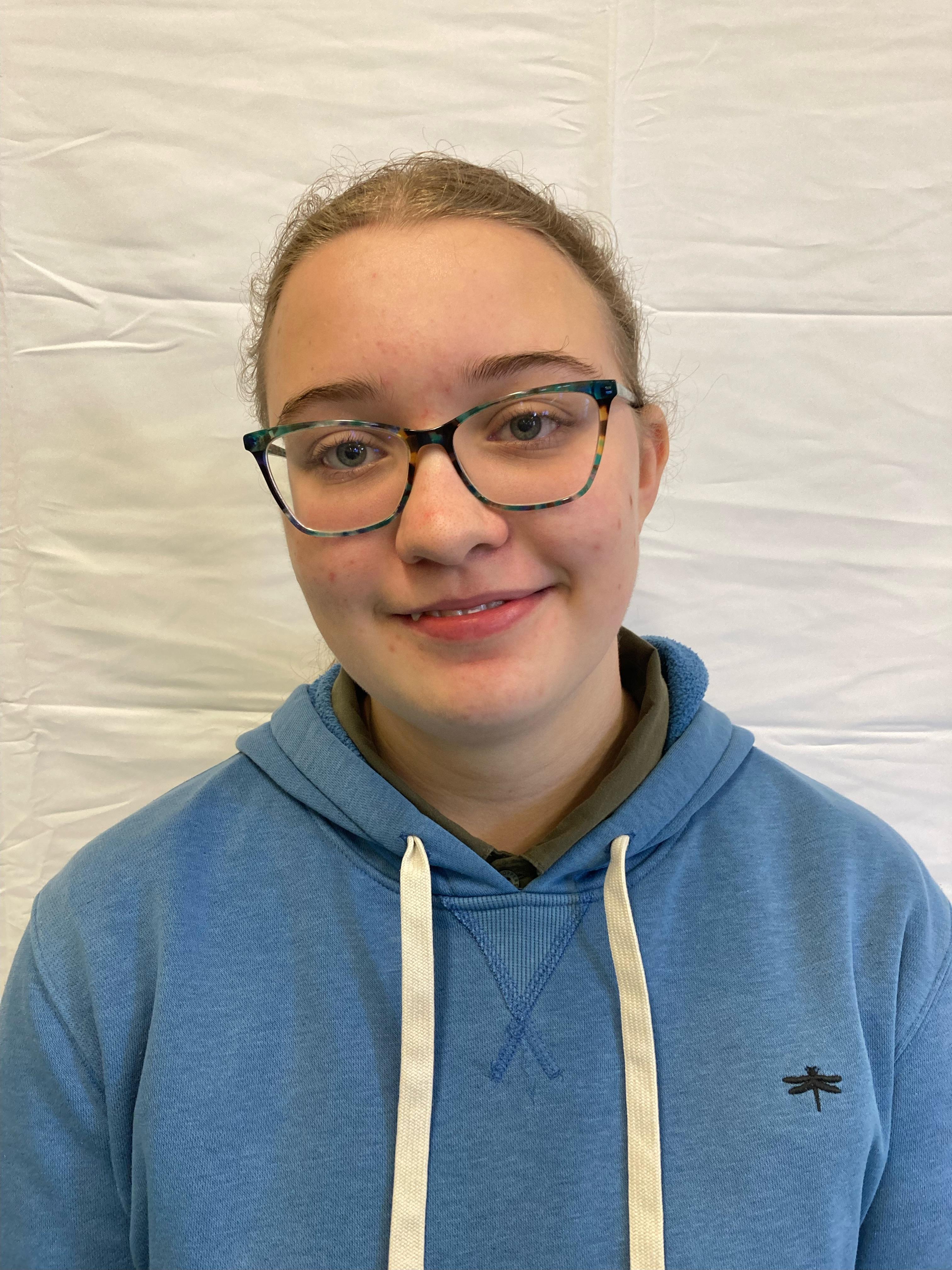 A girl with tied-up brunette hair wearing a blue hoodie and glasses poses for a profile picture.