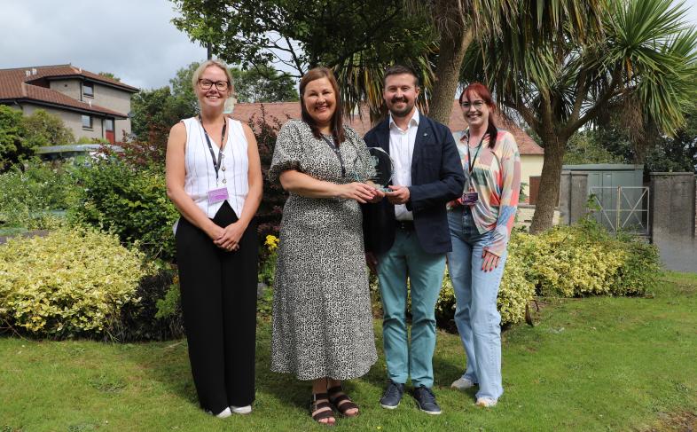 Four people stand in a row in a park. The two people in the centre of the row are Elaine Beswick and Kevin Ward; Kevin is presenting Elaine with the Jane Davies runner up award. 