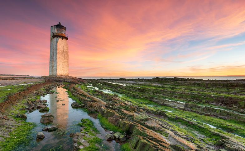 Image shows the lighthouse at Southerness beach pictured against a pink and blue sky, with rocky beach in the foreground