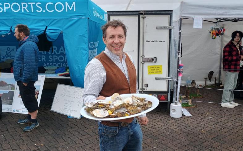 Man in a brown vest holds a platter of oysters, smiling at an outdoor market. Blue tent and other visitors in the background, creating a lively atmosphere.