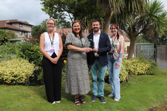 Four people stand in a row in a park. The two people in the centre of the row are Elaine Beswick and Kevin Ward; Kevin is presenting Elaine with the Jane Davies runner up award. 