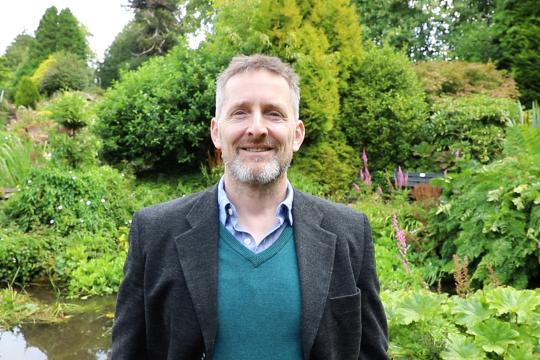 Cllr. Stephen Thompson smiles at the camera, pictured against the Crichton gardens on a bright day.