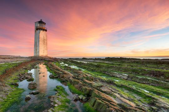 Image shows the lighthouse at Southerness beach pictured against a pink and blue sky, with rocky beach in the foreground