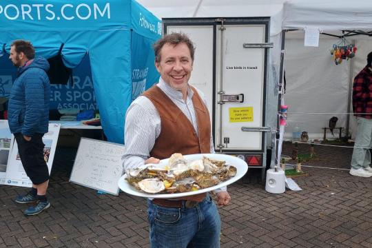 Man in a brown vest holds a platter of oysters, smiling at an outdoor market. Blue tent and other visitors in the background, creating a lively atmosphere.
