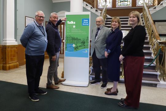 New digital display promoting library talks at Ewart Library, with five people standing alongside.